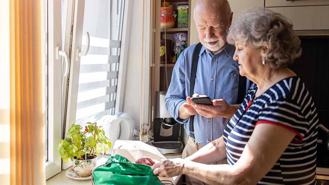 Älteres Ehepaar, Frau packt Einkäufe aus, Mann tippt auf Taschenrechner