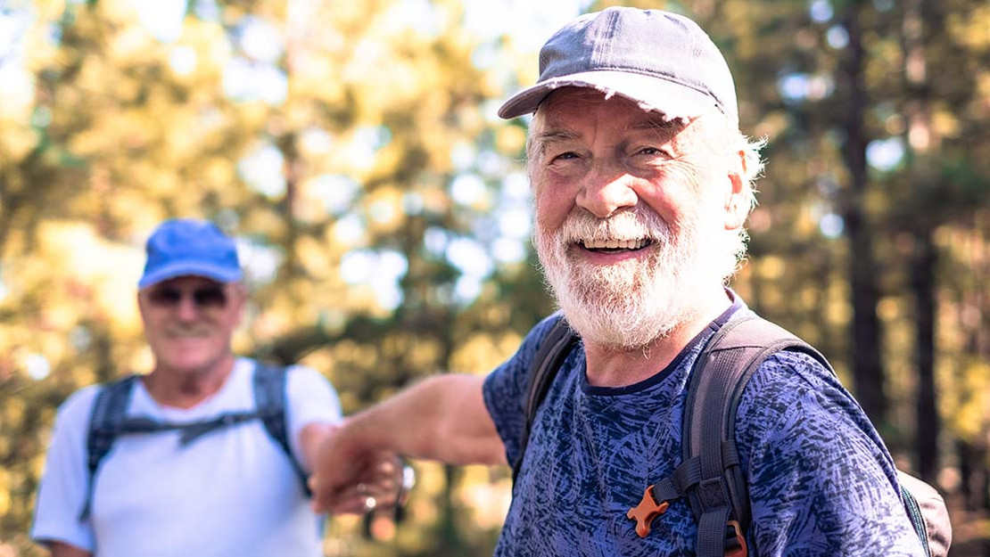 Zwei ältere Männer halten sich an den Händen während sie im Wald wandern. 