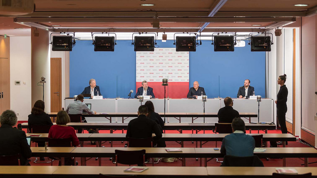 Vier Menschen sitzen auf einem Podium. Journalist*innen im Zuschauerraum. 