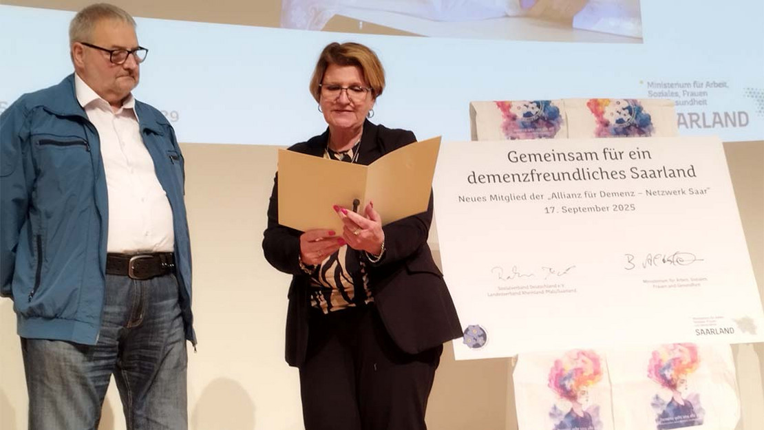 Zwei Personen stehen auf einer Bühne, die Frau hält eine Urkunde in der Hand.