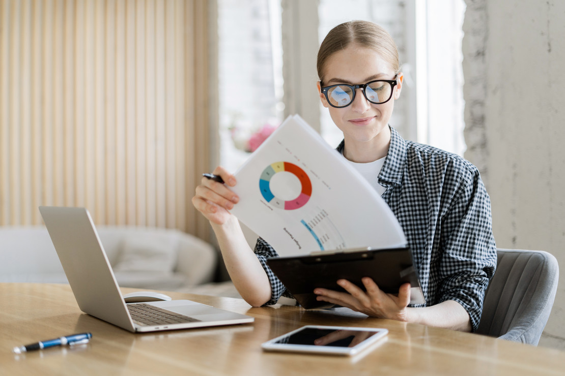 Junge Frau sitzt an Notebook mit Statistiken in der Hand.